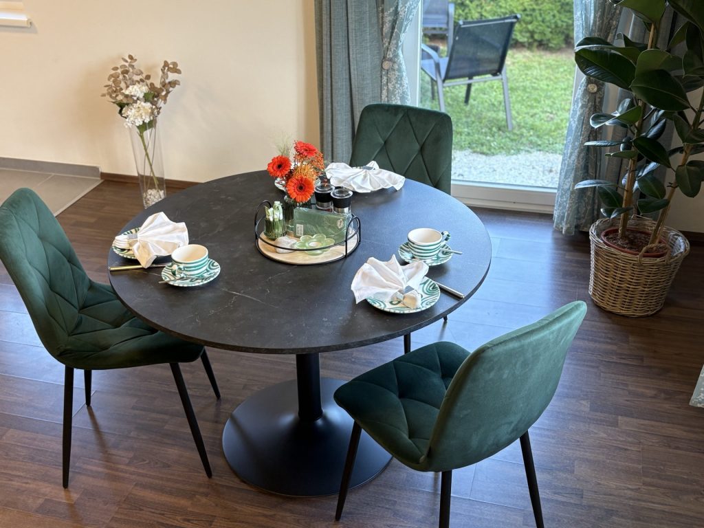 Ein runder schwarzer Tisch für drei Personen mit grünen Stühlen, weißen Servietten, grün gemusterten Tellern, einem Blumengesteck und Blick durch eine Glastür auf eine Terrasse und Grünflächen.
