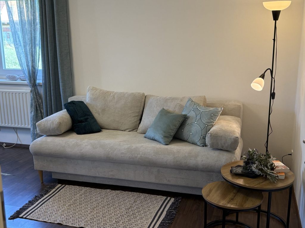 Ein beiges Sofa mit verschiedenen Kissen steht neben einem Fenster mit blauen Vorhängen, neben einem kleinen Tisch und einer Stehlampe in einem gemütlichen Wohnzimmer.